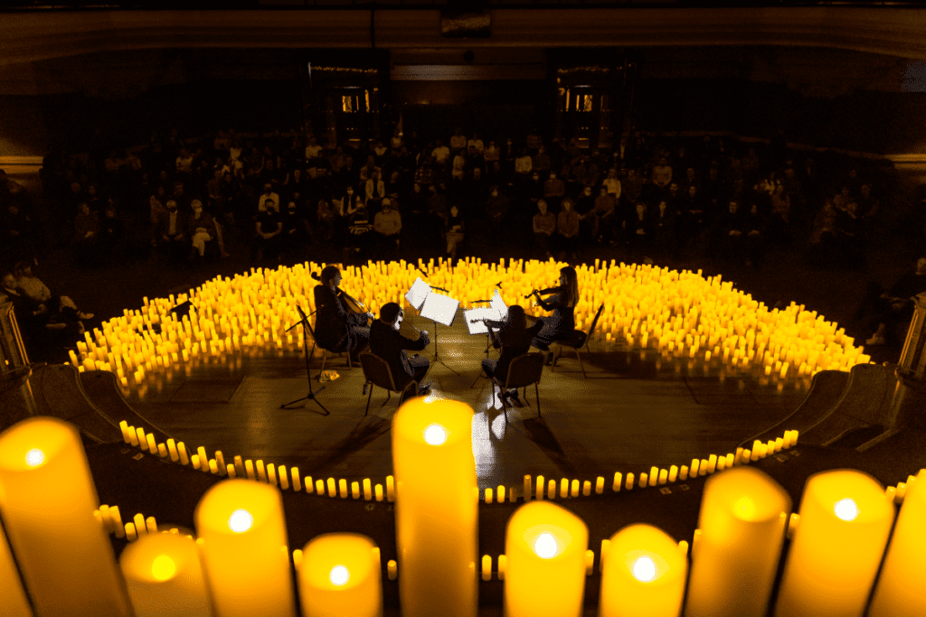 A close up of candles on display at a height with a string quartet performing below on a stage covered in candles for a Candlelight concert.